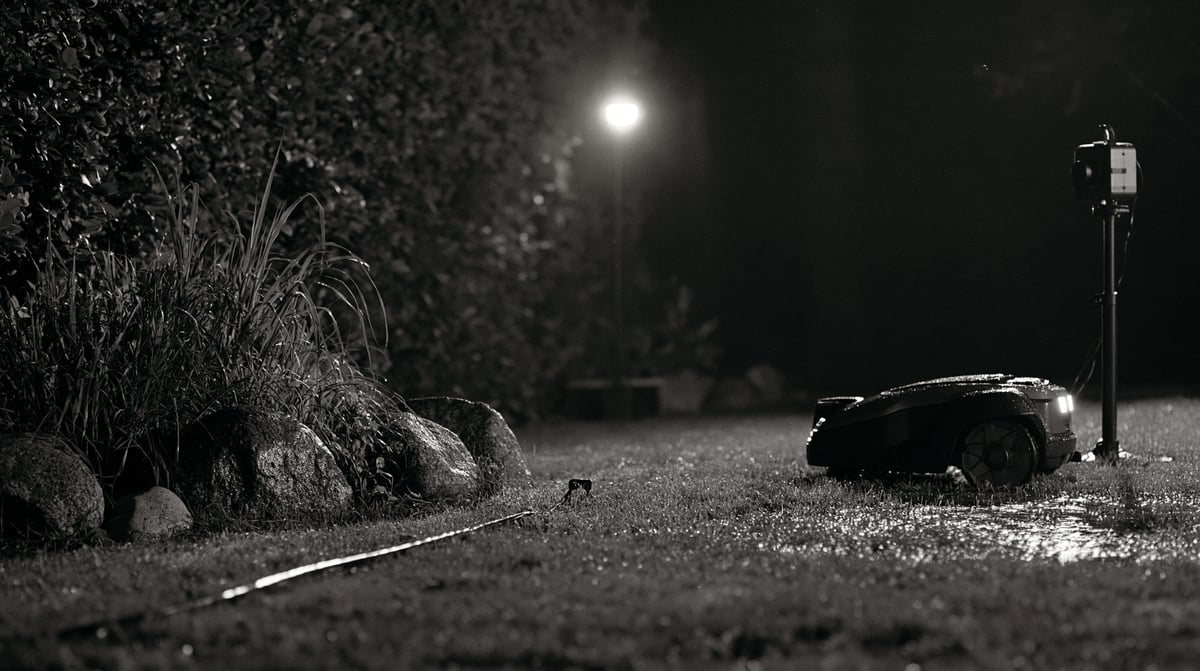 A robotic lawn mower on a wet lawn at night, a fixed camera on a short mast at the border recording it.