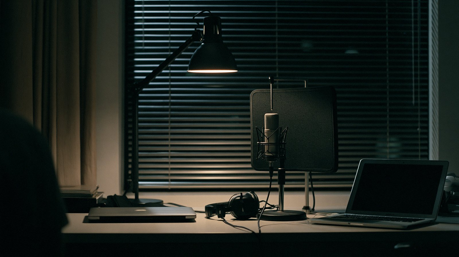 A condenser microphone with a reflection filter on an office desk at night, headphones, a laptop and blinds behind.